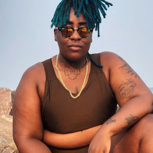 A person wearing sunglasses with blue dreadlocks is crouching down and wearing a dark brown binder